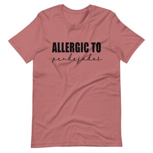 May include: A pink t-shirt with the text "ALLERGIC TO pendejadas" in black.