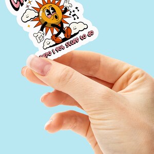 May include: A sticker with a cartoon sun character with a pink, orange, and yellow color scheme. The sun is walking on clouds and has the text "cansada pero I got stuff to do" written below it.