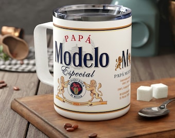 May include: White travel mug with a black lid and handle. The mug features a graphic of the Modelo Especial beer logo with the words "Papá Modelo Especial" in blue and white text. The logo includes a crest with two lions and the words "El mejor papa de Mexico" in a circle.