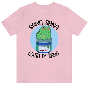 May include: A light pink t-shirt featuring a cartoon frog sitting on a VapoR&uacute; jar. The frog is green with a small bandage. The text "Sana Sana" is arched above the frog, and "Colita de Rana" is below.