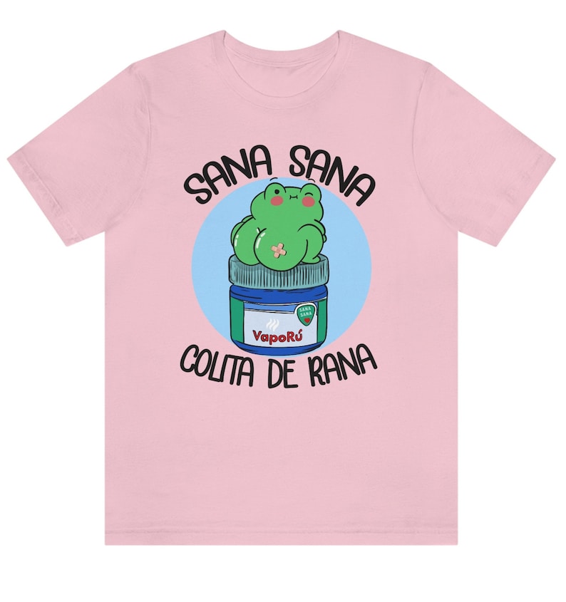 May include: A light pink t-shirt featuring a cartoon frog sitting on a VapoR&uacute; jar. The frog is green with a small bandage. The text "Sana Sana" is arched above the frog, and "Colita de Rana" is below.