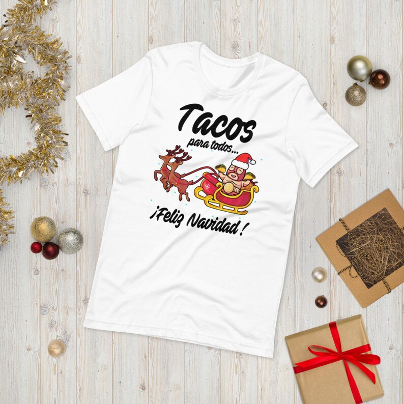 May include: White t-shirt with a festive graphic featuring a taco character in a Santa hat riding a sleigh pulled by reindeer. The text reads "Tacos para todos... &iexcl;Feliz Navidad!"