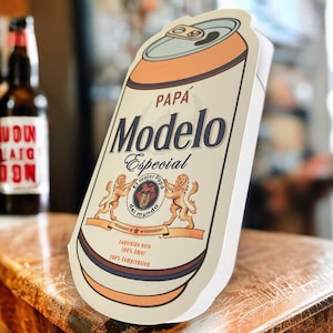 May include: A white and orange can with a brown label. The label has the text "Modelo Especial" in a brown font with the words "Pap&aacute;" above it. The label also has a crest with a lion and the text "El Mejor  Fondo del Mundo".