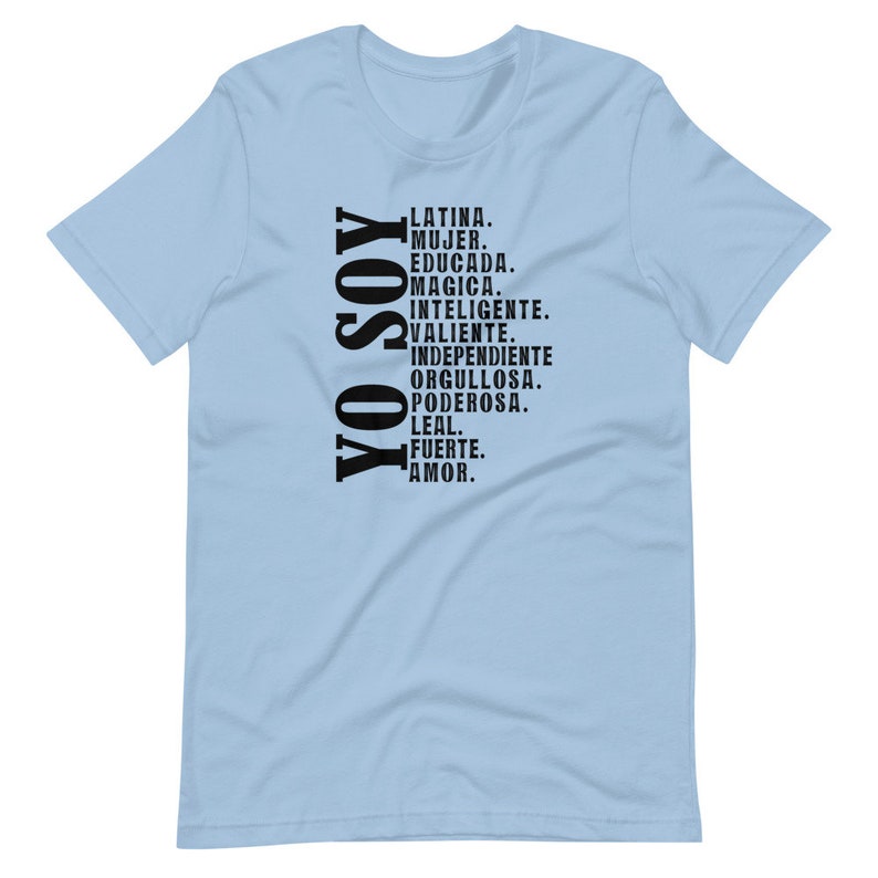 Puede incluir: Camiseta azul claro con texto negro que dice "YO SOY" y una lista de palabras en espa&ntilde;ol que describen a una mujer latina. Las palabras incluyen "Latina, Mujer, Educada, Magica, Inteligente, Valiente, Independiente, Orgullosa, Poderosa, Leal, Fuerte, Amor."