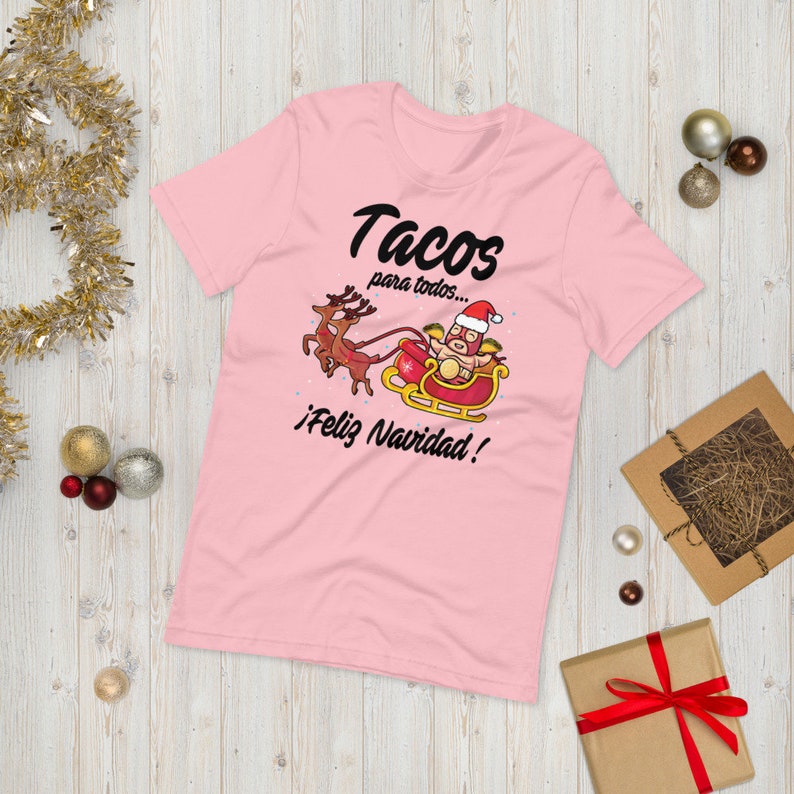 May include: A light pink t-shirt with the text "Tacos para todos... &iexcl;Feliz Navidad!" and a graphic of Santa Claus in a taco costume riding a sleigh pulled by reindeer. The shirt is a festive holiday design.
