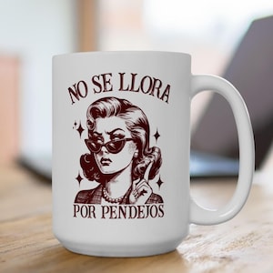 May include: White ceramic mug featuring a retro illustration of a woman wearing sunglasses, with the text "NO SE LLORA POR PENDEJOS" in maroon. The mug has a curved handle and is set against a blurred background.