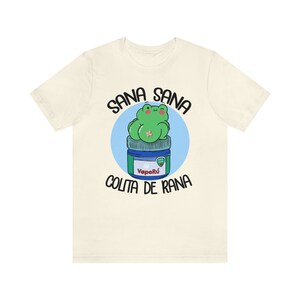 May include: Cream-colored t-shirt featuring a cartoon frog on a blue and green jar. The text "SANA SANA COLITA DE RANA" is arched above and below the frog illustration. The jar has the text "Vapor&uacute;" on it.