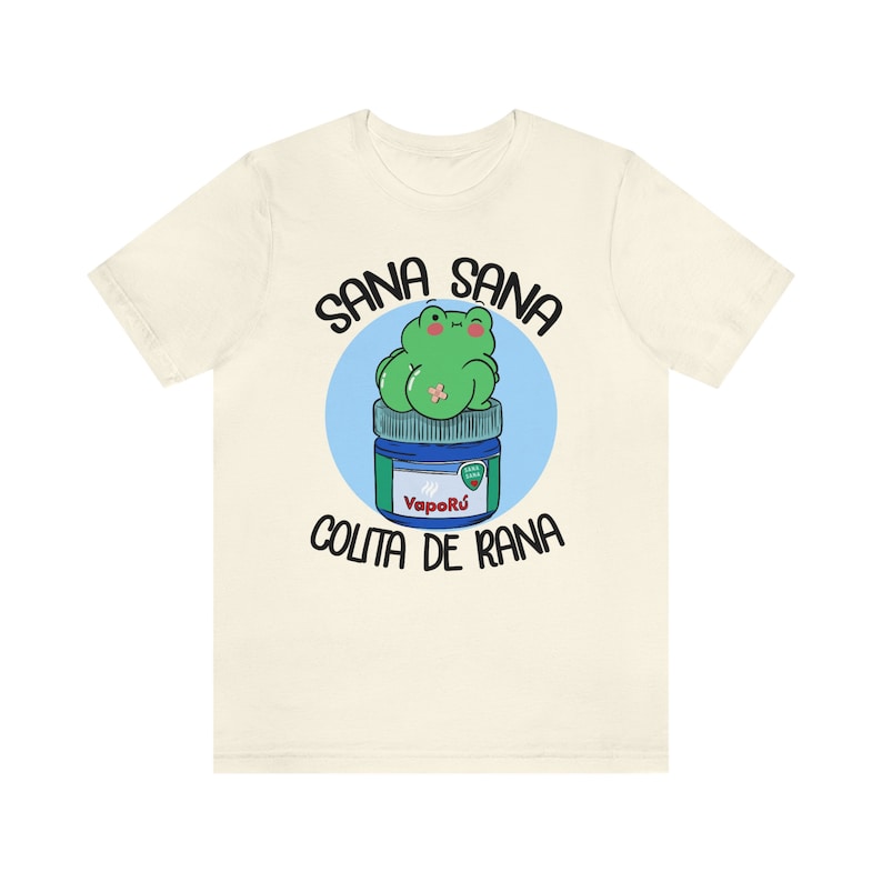 May include: Cream-colored t-shirt featuring a cartoon frog on a blue and green jar. The text "SANA SANA COLITA DE RANA" is arched above and below the frog illustration. The jar has the text "Vapor&uacute;" on it.