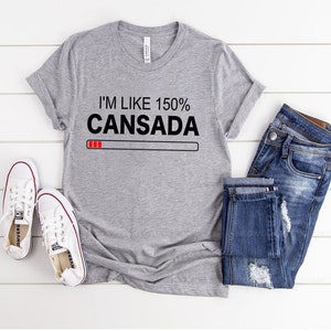 May include: Gray t-shirt with the text "I'm like 150% Cansada" and a red progress bar underneath. The shirt is laid flat on a white wooden surface with a pair of white sneakers and blue jeans.
