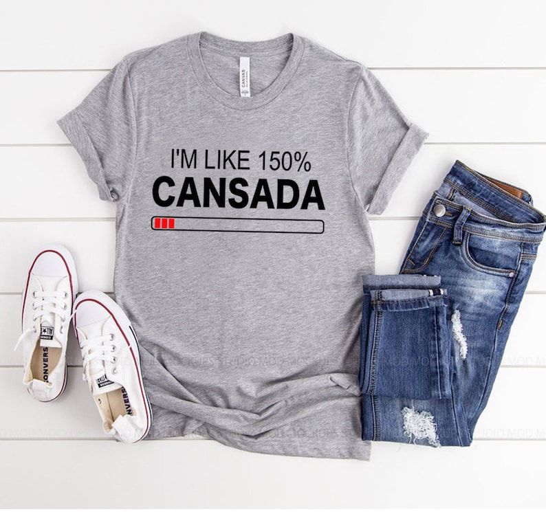 May include: Gray t-shirt with the text "I'm like 150% Cansada" and a red progress bar underneath. The shirt is laid flat on a white wooden surface with a pair of white sneakers and blue jeans.