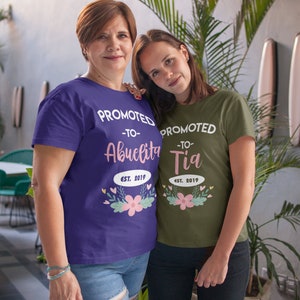 May include: Two women wearing matching t-shirts. One shirt is purple and says "PROMOTED -TO- Abuelita EST. 2019" with floral accents. The other shirt is olive green and says "PROMOTED -TO- Tia EST. 2019" with floral accents.