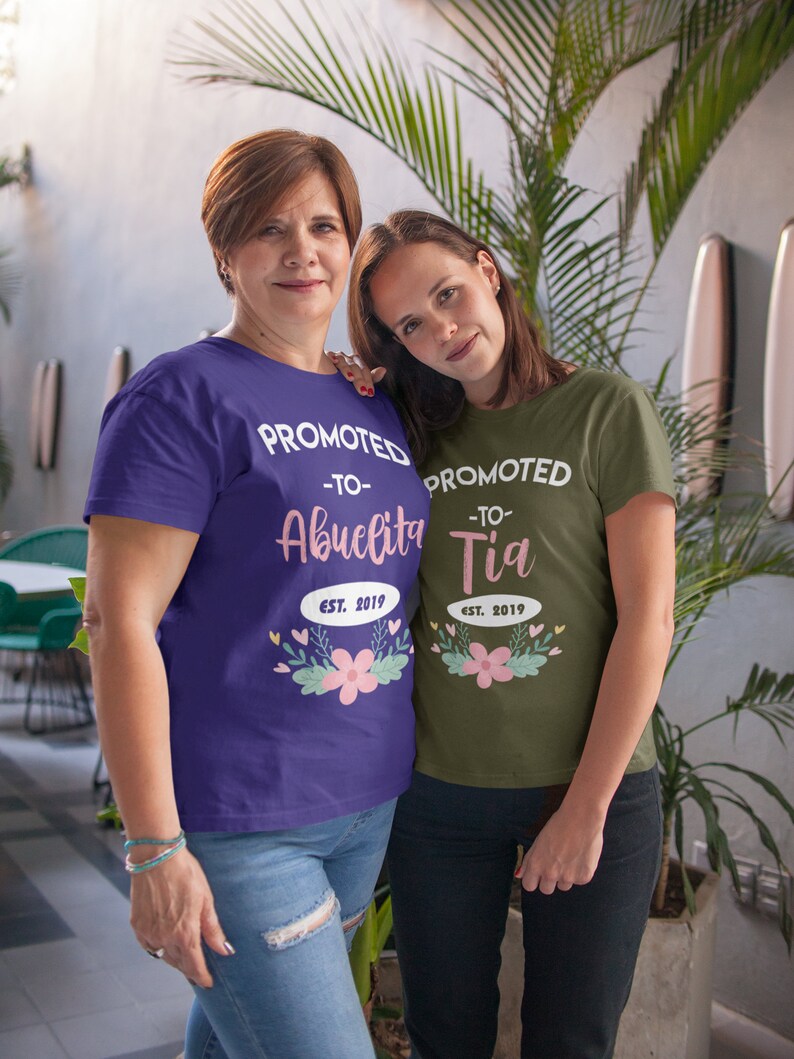 May include: Two women wearing matching t-shirts. One shirt is purple and says "PROMOTED -TO- Abuelita EST. 2019" with floral accents. The other shirt is olive green and says "PROMOTED -TO- Tia EST. 2019" with floral accents.
