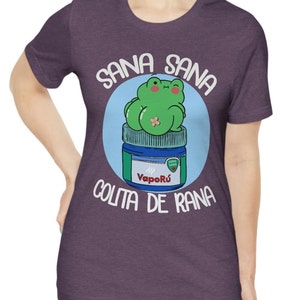 May include: Purple t-shirt featuring a cartoon frog sitting on a blue and green jar. The text "SANA SANA COLITA DE RANA" is printed around the frog. The frog is green with pink cheeks and a bandage. The jar has the word "Vapor&uacute;" on it.