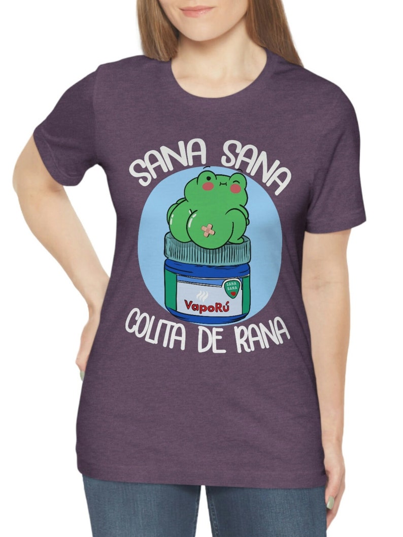 May include: Purple t-shirt featuring a cartoon frog sitting on a blue and green jar. The text "SANA SANA COLITA DE RANA" is printed around the frog. The frog is green with pink cheeks and a bandage. The jar has the word "Vapor&uacute;" on it.
