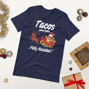 May include: Navy blue t-shirt with a festive graphic. The design features a taco character in a Santa hat riding a sleigh pulled by reindeer, with the words "Tacos para todos... &iexcl;Feliz Navidad!" in white.