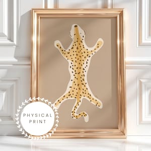 May include: A framed wall art print featuring a stylised, flat illustration of a leopard skin rug in shades of tan, cream, and dark brown spots. The artwork is encased in a metallic gold frame, displaying a centred, symmetrical animal hide design.