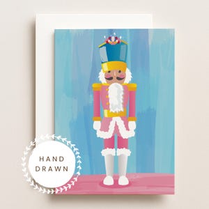 Peut inclure: Une carte de vœux présentant une illustration numérique d'un soldat casse-noisette en uniforme rose avec des bordures en fourrure blanche, un chapeau bleu à détails dorés et une barbe blanche. Le texte "HAND DRAWN" est inscrit dans un emblème.