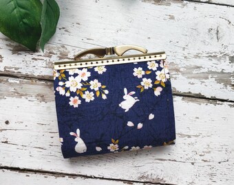 Puede incluir: Una pequeña cartera de tela azul con estampado floral y conejos blancos. La cartera tiene un cierre de metal dorado.