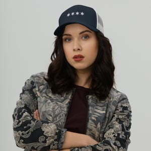 May include: A woman wearing a navy blue and white trucker hat with five white stars on the front. She is wearing a black and white floral patterned jacket.