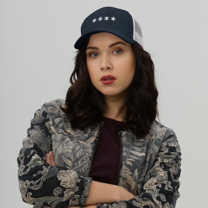 May include: A woman wearing a navy blue and white trucker hat with five white stars on the front. She is wearing a black and white floral patterned jacket.