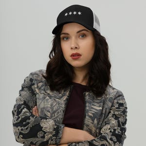 May include: A woman wearing a black and white trucker hat with five white stars on the front. She is wearing a black and white patterned jacket and has long black hair.