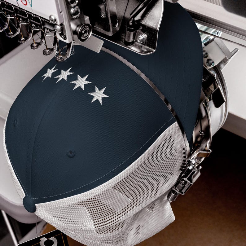 May include: A navy blue and white baseball cap with a mesh back is being embroidered with five white stars. The cap is being stitched on a commercial embroidery machine.