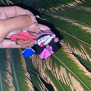May include: A pair of dangle earrings featuring cartoon characters from the animated television series, The Powerpuff Girls. The earrings are made of metal and have a gold finish. The left earring features a pink-haired character with a pink jacket and a red hat. The right earring features a black-haired character with a pink jacket and a red hat.