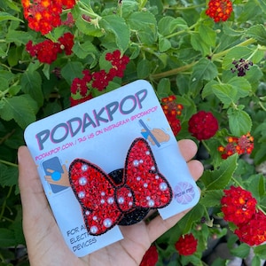 May include: A red bedazzled bow-shaped phone grip with white pearl accents. The grip is on a black base and is held in front of a background of red flowers and green leaves. The product is from PodakPop.