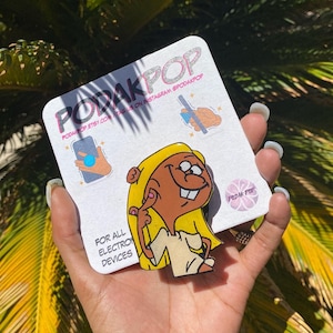 Könnte beinhalten: Ein Telefonhalter in Form einer Zeichentrickfigur mit gelbem Haar und einem gebräunten Teint. Der Halter befindet sich auf einer weißen Rückseite mit dem Text "PODAKPOP" und "FOR ALL ELECTRONIC DEVICES". Der Hintergrund ist verschwommen grün.