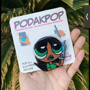May include: A black and green cartoon character phone grip with the text "PODAKPOP" and "FOR ALL ELECTRONIC DEVICES". The grip is attached to a white card with illustrations and the Etsy shop's website.