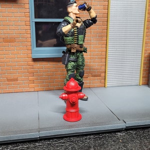 May include: An action figure in military-style camouflage fatigues drinks from a blue can. The figure stands next to a bright red fire hydrant on a gray sidewalk, with a brick wall and a closed metal door in the background.