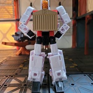May include: A white and red action figure with a tan chest and a faceplate. The figure has a rectangular chest with vertical lines and the letter "R". The figure is standing on a gray surface.