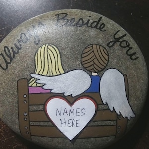 May include: Hand-painted stone with the words "Always Beside You." The artwork depicts two figures with angel wings, seated on a bench. A heart-shaped space is labeled "NAMES HERE." The figures have blonde and brown hair.