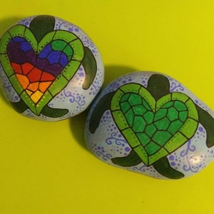 May include: Two painted rocks shaped like sea turtles with hearts on their shells. One heart is rainbow colored and the other is green. Both rocks have a blue background with white dots and swirls.