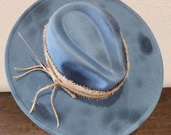 Może przedstawiać: Niebieski filcowy kapelusz fedora z szerokim rondem i brązową wstążką z juty. Kapelusz ma postarzony wygląd z wyblakłym niebieskim kolorem i strzępioną wstążką z juty.