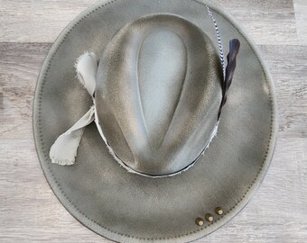 May include: A light gray felt cowboy hat with a white ribbon and a feather accent. The hat has a distressed look and four brass studs around the brim.
