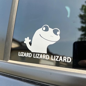 May include: White vinyl sticker featuring a cartoon lizard head and the text "LIZARD LIZARD LIZARD". The sticker is applied to a car window, with a blurred outdoor scene in the background.