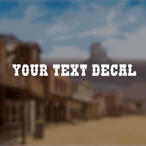 May include: White text decal with the words "YOUR TEXT DECAL" in a western-style font. The background is a blurred image of a western town with a blue sky and mountains.