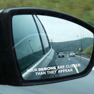 May include: A car's wing mirror reflecting a motorway scene with the text "YOUR DEMONS ARE CLOSER THAN THEY APPEAR" in white. The image shows other vehicles and a distant mountainous landscape.