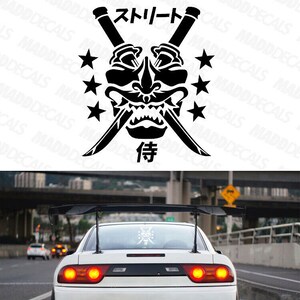 May include: Black graphic featuring a samurai mask, crossed swords, stars, and Japanese text. The design is on the rear window of a white sports car with a spoiler. The car is on a road with other vehicles.