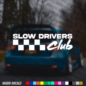 Peut inclure: Autocollant en vinyle blanc avec le texte "SLOW DRIVERS Club" au-dessus d'un motif de drapeau à damier. L'autocollant est affiché à l'arrière d'une voiture bleue. La marque "MADD DECALS" est en bas.