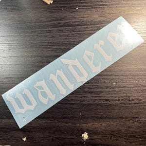 May include: A white vinyl sticker with the word "Wanderer" in a gothic font. The sticker is rectangular and has a light blue backing. The sticker is placed on a dark wooden surface.