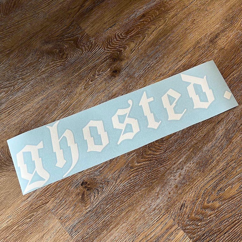 以下が含まれることがあります： 白いゴシック体で「ghosted」という文字が書かれた、水色の長方形のステッカー。木製の表面に貼られています。
