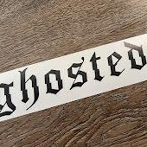 以下が含まれることがあります： 黒いゴシック体で「ghosted」と書かれた白い長方形のステッカー。ステッカーは木製の表面にあります。