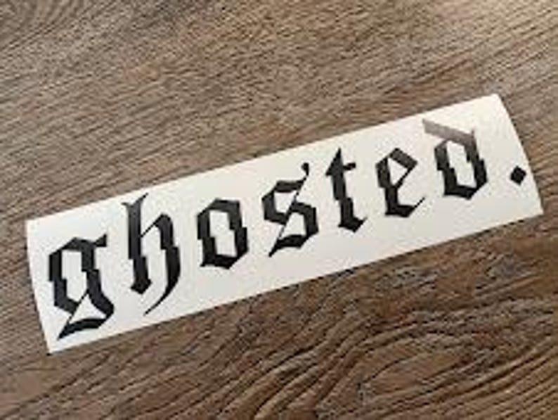 以下が含まれることがあります： 黒いゴシック体で「ghosted」と書かれた白い長方形のステッカー。ステッカーは木製の表面にあります。
