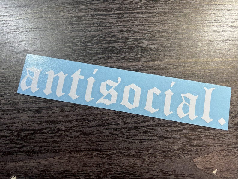 May include: A light blue sticker with the word "antisocial" in a white gothic font. The sticker is rectangular and has a glossy finish. The word is centered and takes up most of the sticker's surface.