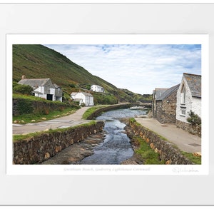 May include: A scenic view of a small village in Cornwall, England. The village is built on a hillside overlooking a small river. The houses are white with stone walls and slate roofs. The river flows through the village and out to the sea. The sky is blue with white clouds. The image is captioned "Treen, Beach, Godrevy Lighthouse, Cornwall".