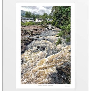 May include: A photograph of the Falls of Dochart in Scotland. The rushing water flows over rocks and through a narrow gorge.  The falls are surrounded by lush green trees and a small village in the distance.