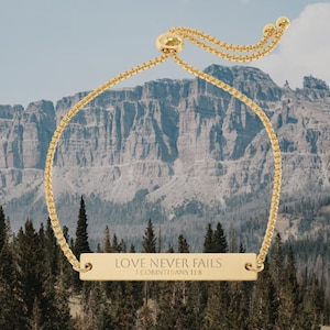 Peut inclure: Bracelet doré avec un pendentif rectangulaire. Le pendentif porte l'inscription "LOVE NEVER FAILS" et "1 CORINTHIANS 13:8". Le bracelet est présenté sur un fond de montagnes et de conifères.