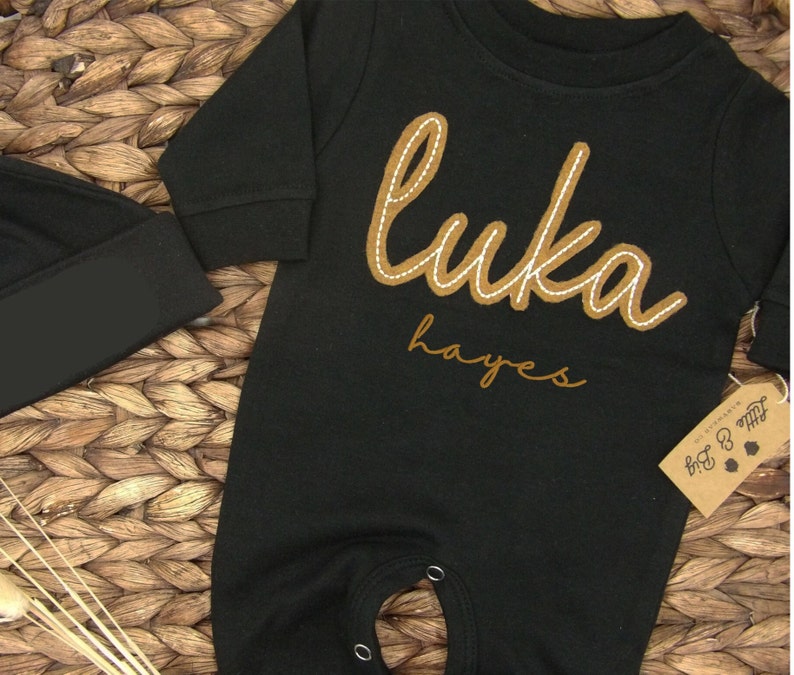 May include: Black baby onesie with brown embroidered lettering that says "Luka Hayes".